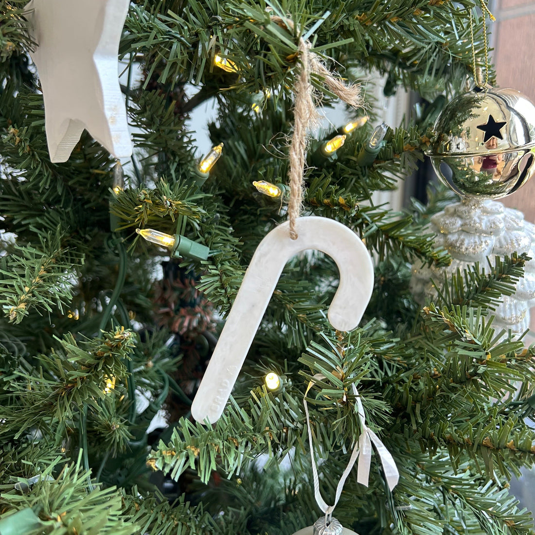 A white recycled plastic candy cane decoration hanging on a green Christmas tree with warm string lights, next to a star decoration.