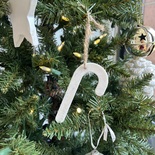 A white recycled plastic candy cane decoration hanging on a green Christmas tree with warm string lights, next to a star decoration.